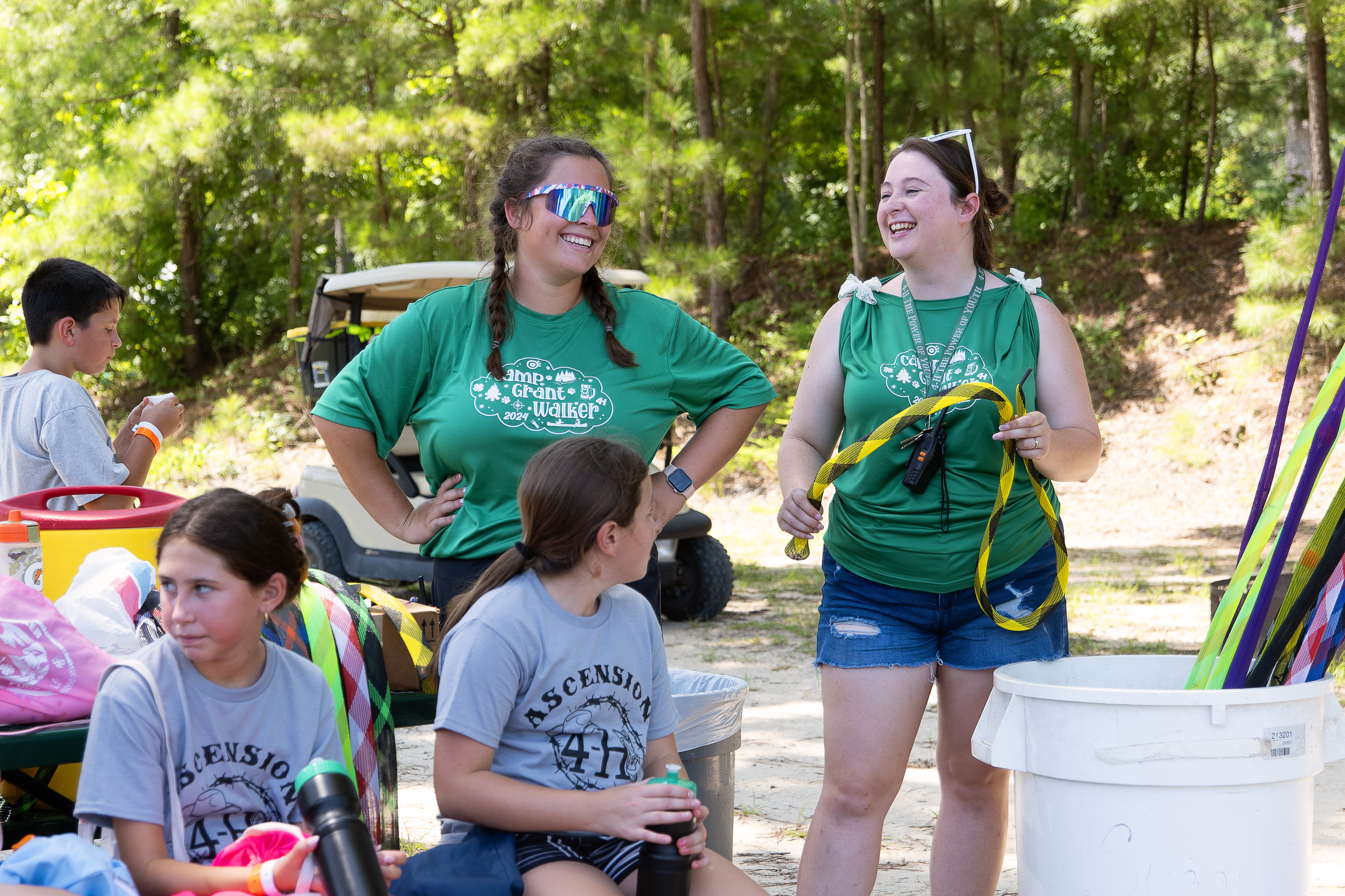 Two women laugh while standing near children seated at picnic table.
